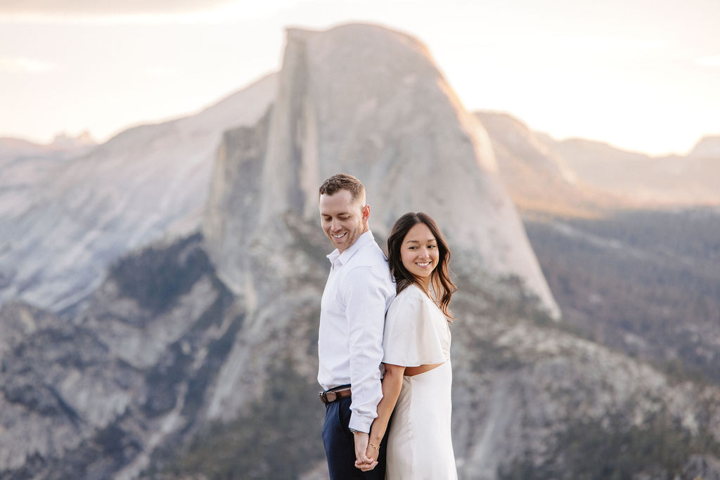 A couple stands on a rock ledge, gazing at Half Dome in Yosemite National Park, with a sunset sky in the background for their yosemite engagement photos