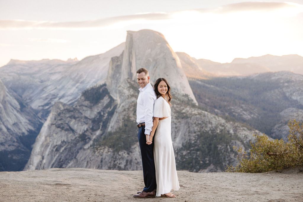 A couple stands on a rock ledge, gazing at Half Dome in Yosemite National Park, with a sunset sky in the background for their yosemite engagement photos