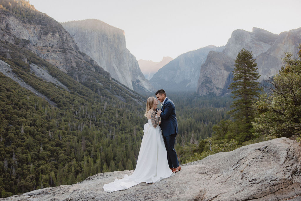 A bride and groom stand on a rocky ledge with a scenic view of a forested valley and mountains in the background for an elopement at bridalveil falls