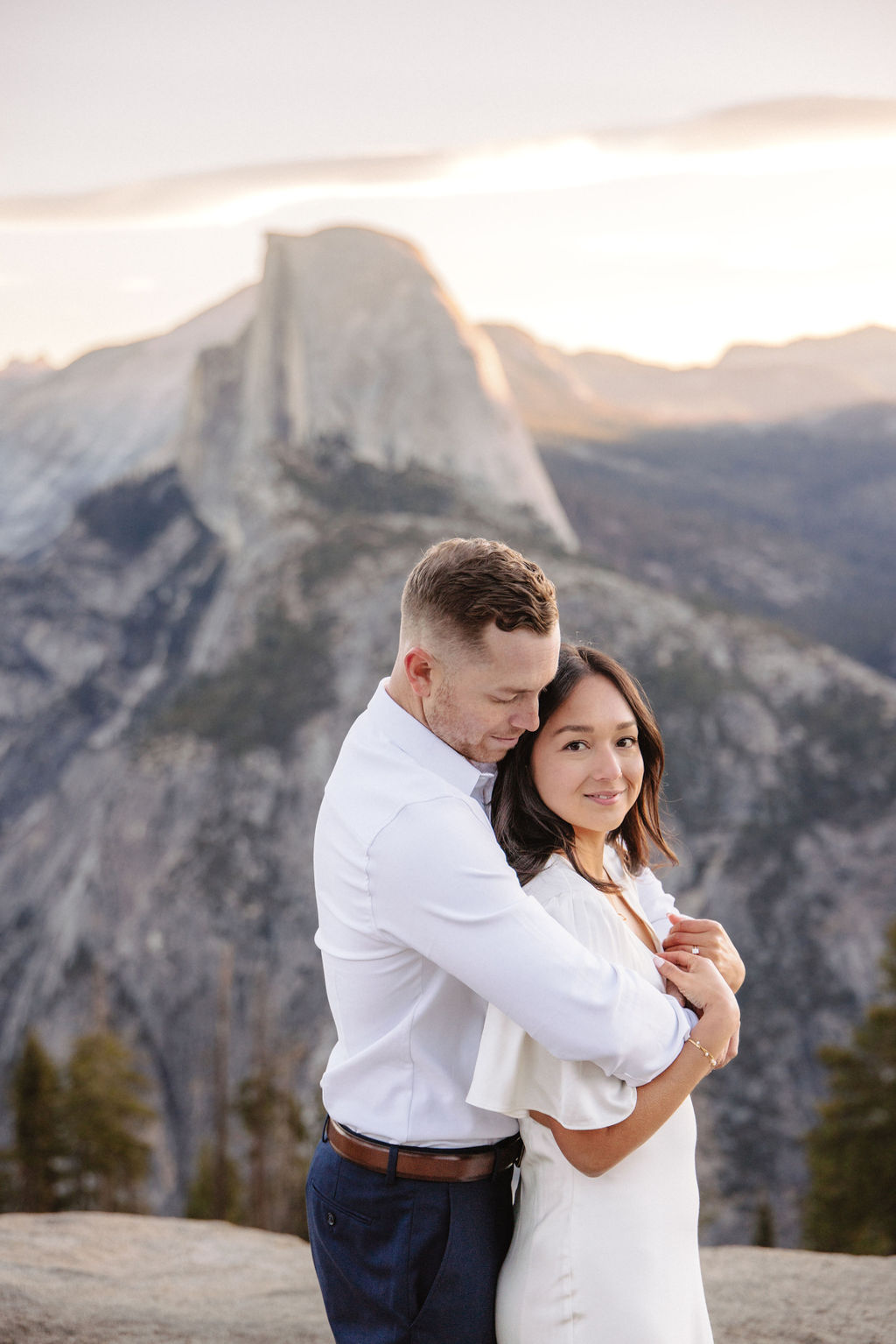 A couple stands on a rock ledge, gazing at Half Dome in Yosemite National Park, with a sunset sky in the background for their yosemite engagement photos