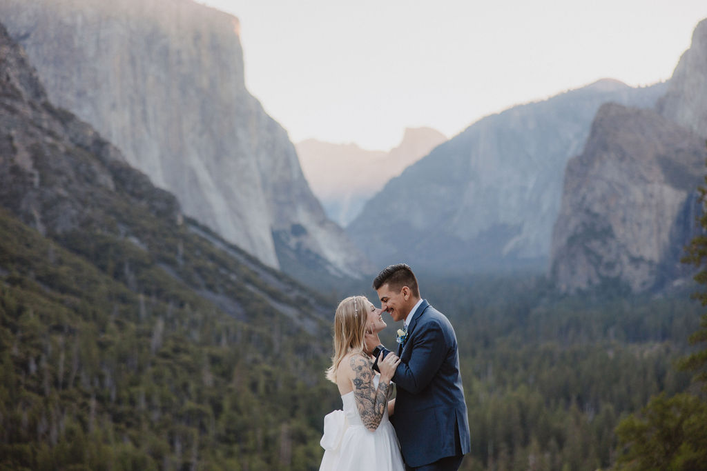 A bride and groom stand on a rocky ledge with a scenic view of a forested valley and mountains in the background for an elopement at bridalveil falls