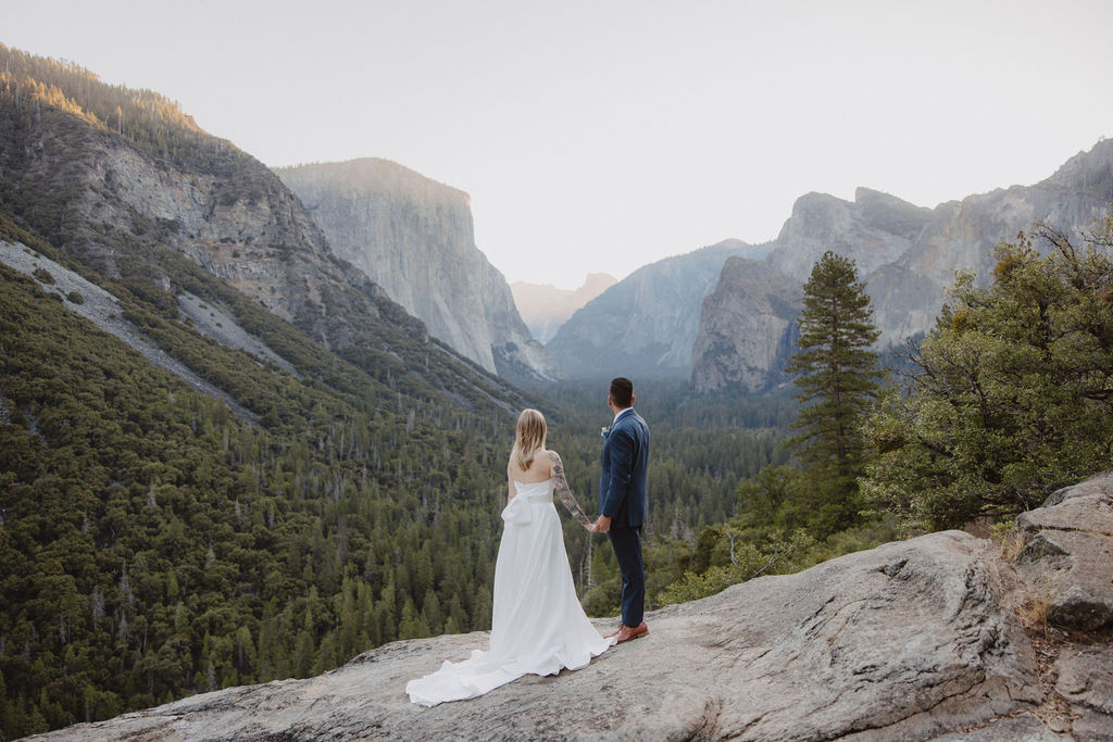 A bride and groom stand on a rocky cliff in wedding attire, with a forest and mountains in the background for an elopement at bridalveil falls