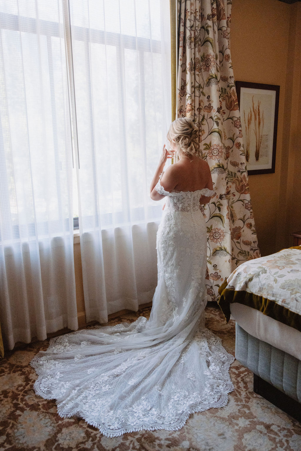 A woman in a white lace strapless dress stands by a window with floral curtains, looking outside.