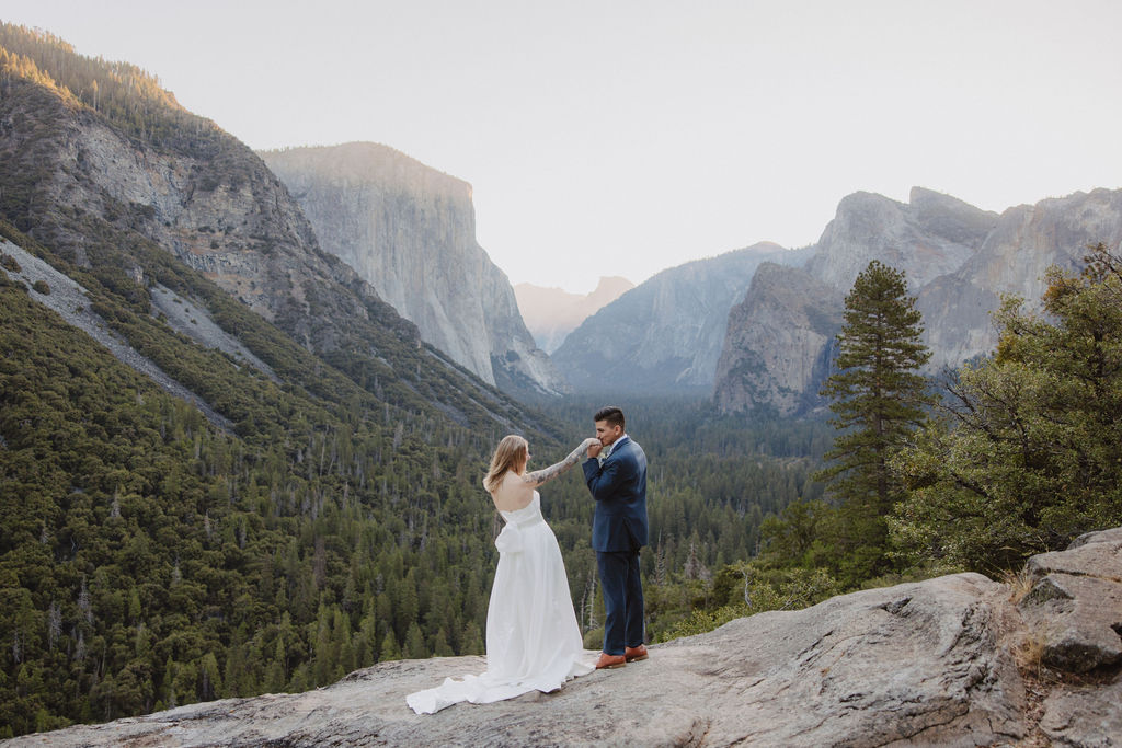 A bride and groom stand on a rocky cliff in wedding attire, with a forest and mountains in the background for an elopement at bridalveil falls