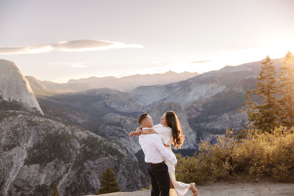 A couple stands on a rock ledge, gazing at Half Dome in Yosemite National Park, with a sunset sky in the background for their yosemite engagement photos