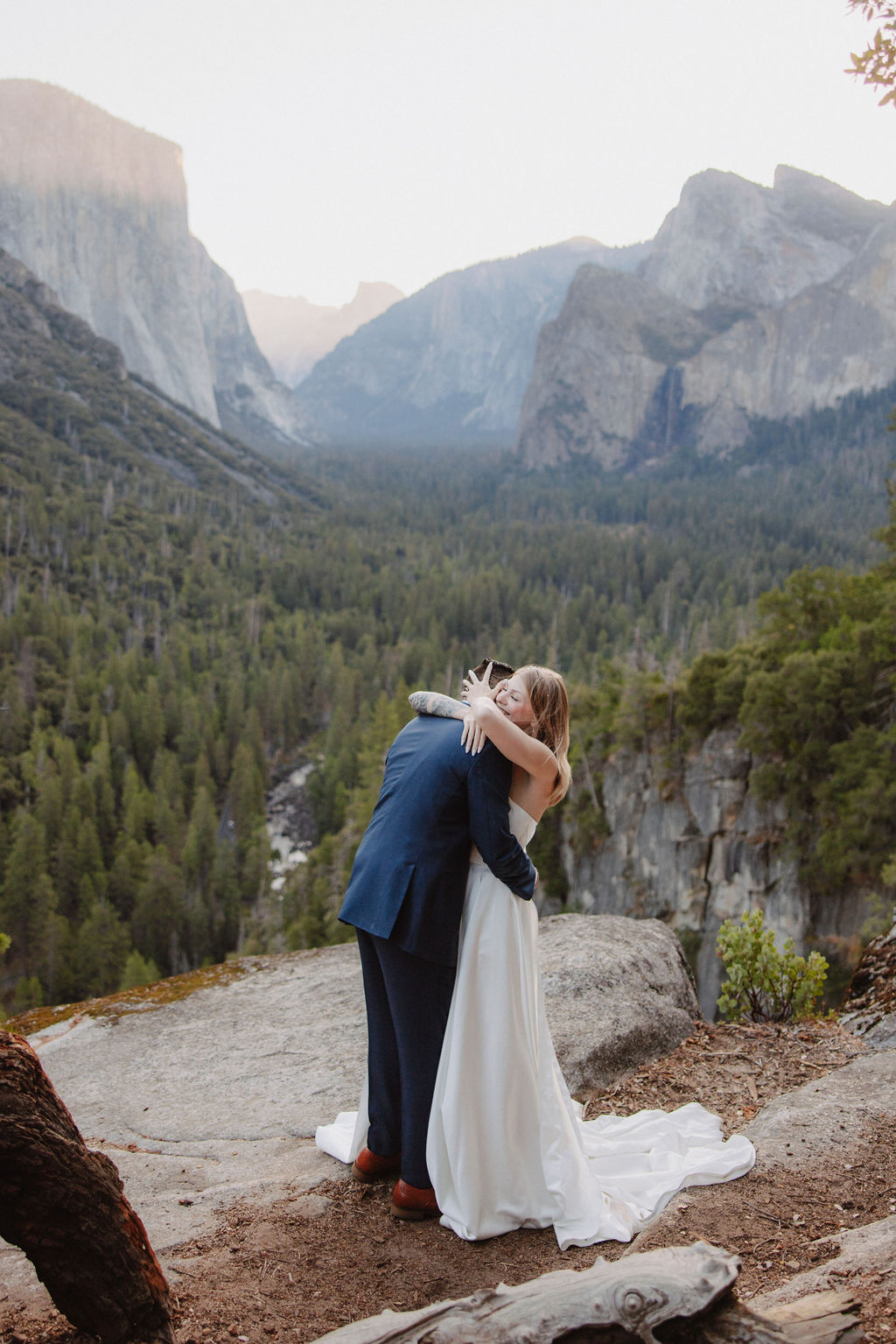 A bride and groom stand on a rocky ledge with a scenic view of a forested valley and mountains in the background for an elopement at bridalveil falls