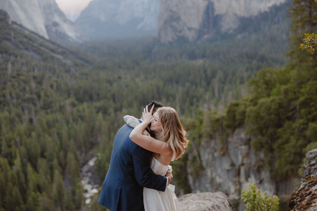 A bride and groom stand on a rocky ledge with a scenic view of a forested valley and mountains in the background for an elopement at bridalveil falls