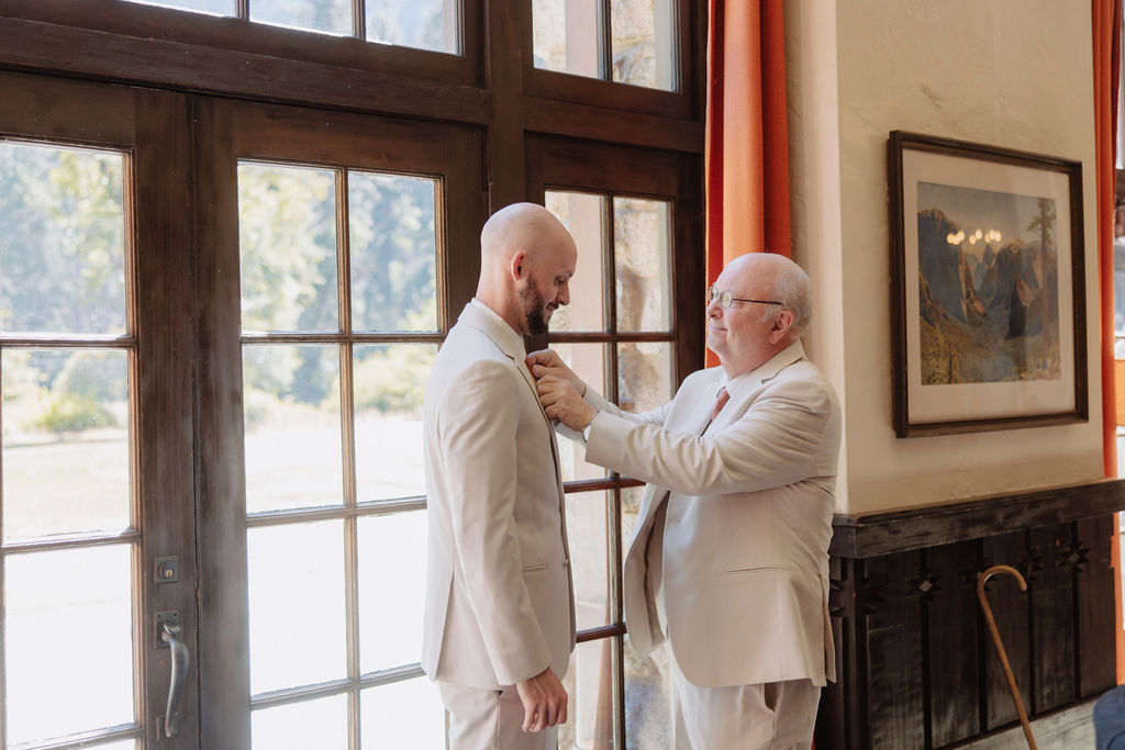 Two men in suits stand near large windows; an older man adjusts the younger man's tie.