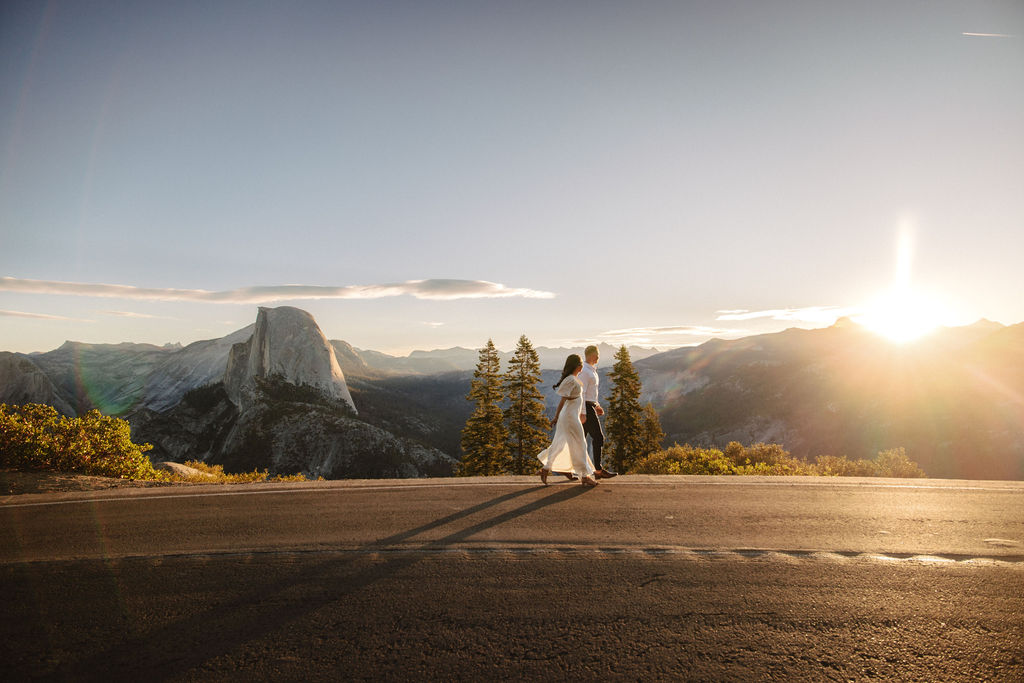 A couple walks hand in hand along a road with Half Dome and the setting sun in the background.