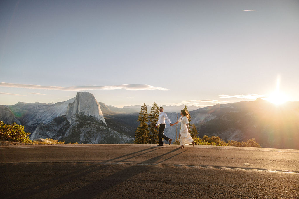 A couple walks hand in hand along a road with Half Dome and the setting sun in the background.