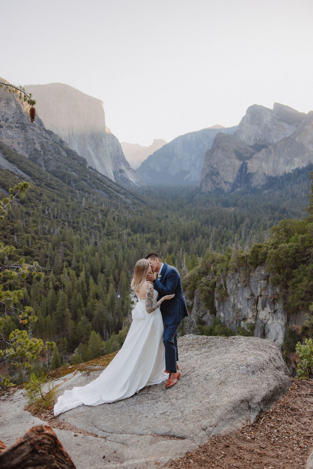 A bride and groom stand on a rocky ledge with a scenic view of a forested valley and mountains in the background for an elopement at bridalveil falls