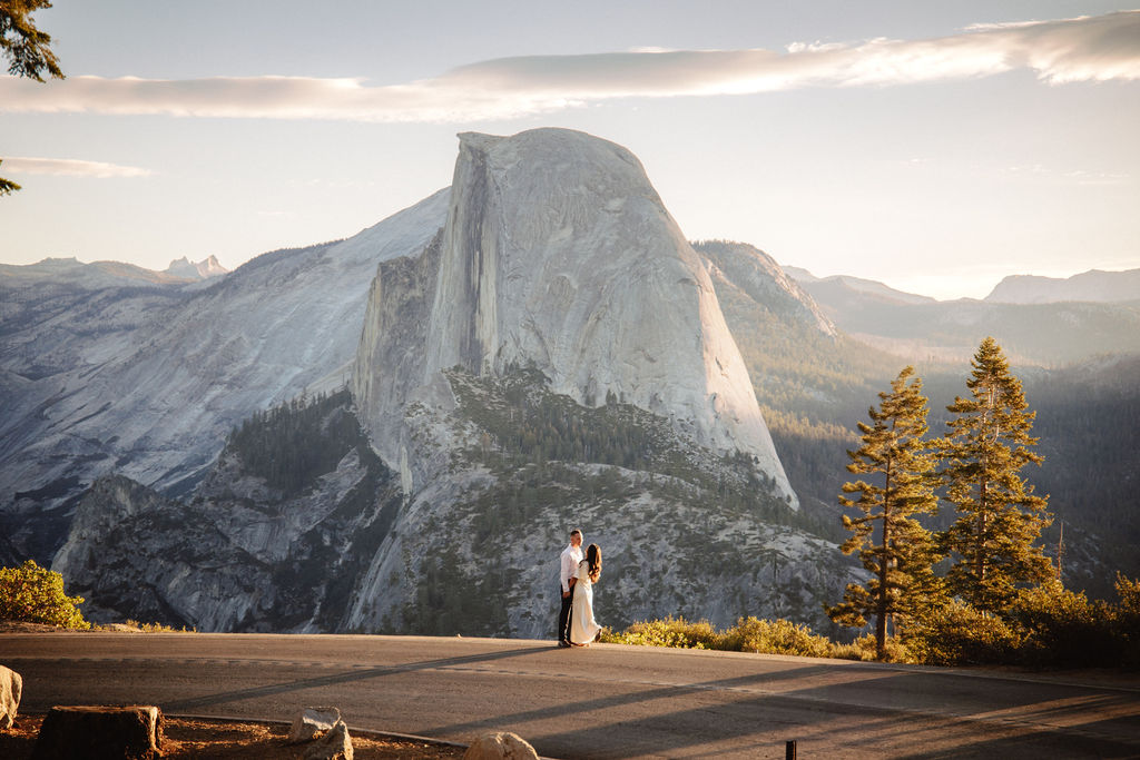 A couple walks hand in hand along a road with Half Dome and the setting sun in the background.