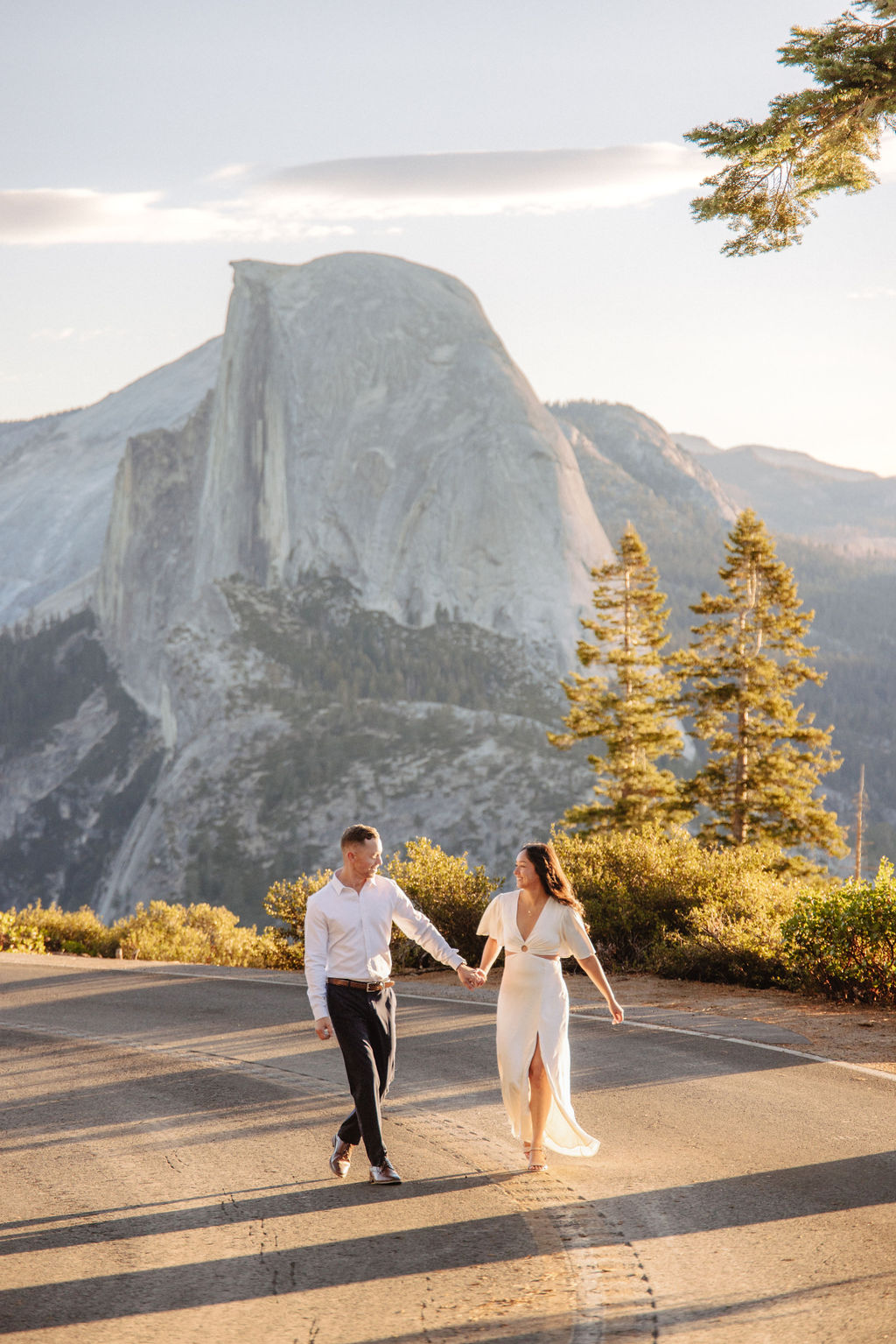 A couple walks hand in hand along a road with Half Dome and the setting sun in the background.