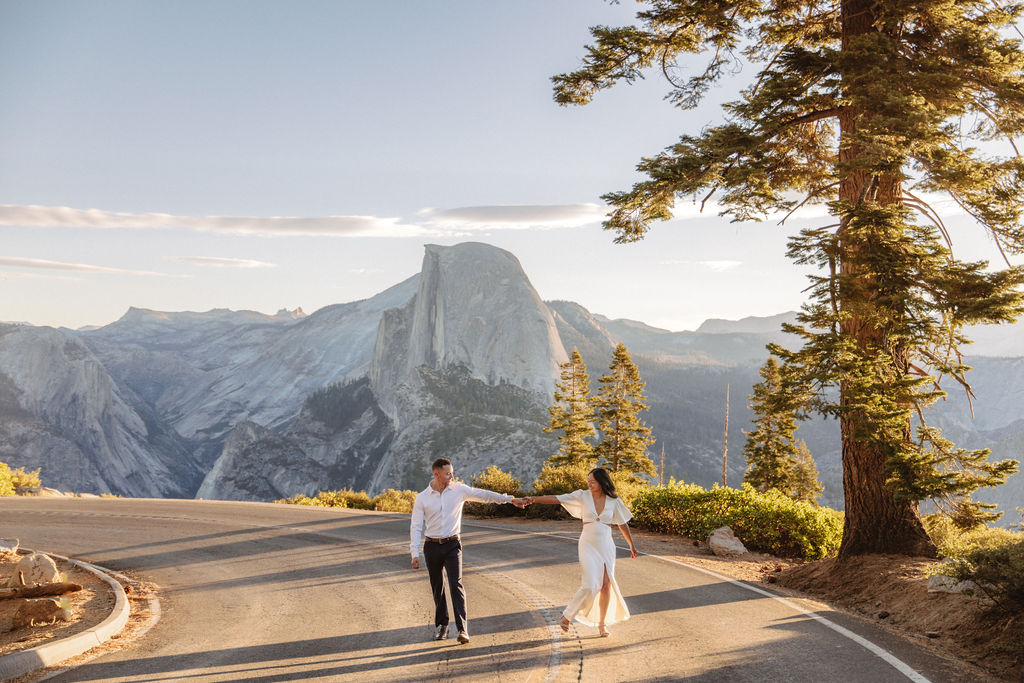 A couple walks hand in hand along a road with Half Dome and the setting sun in the background.