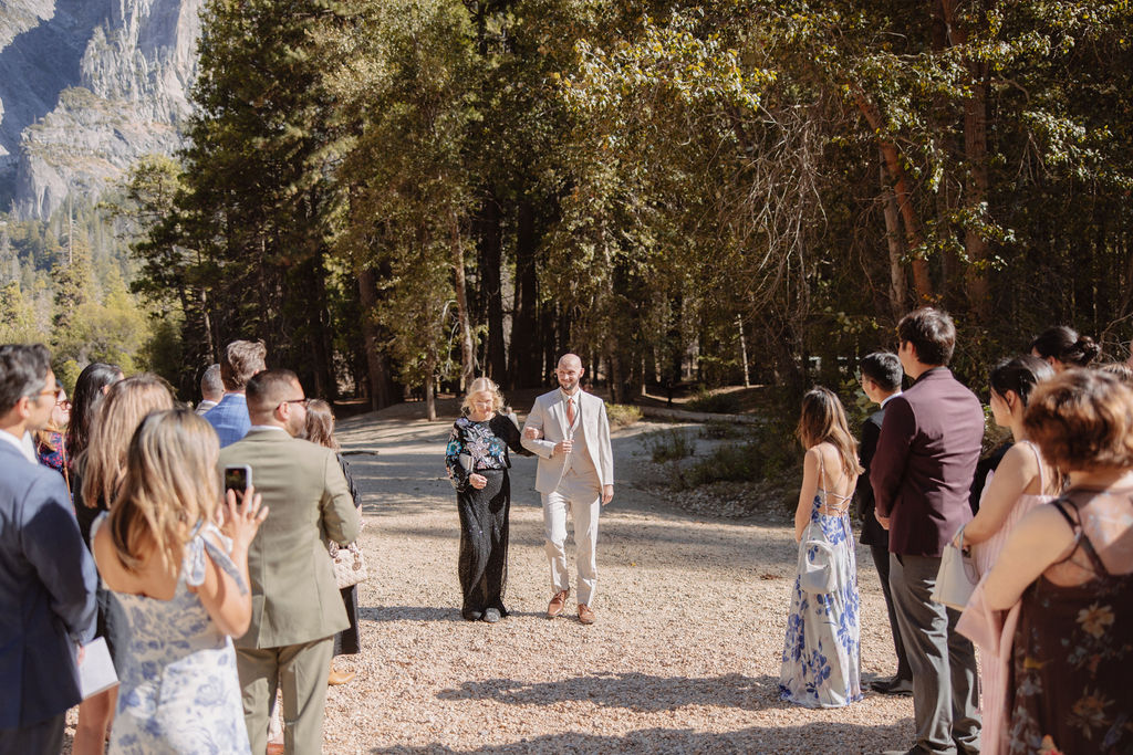 A couple walks hand in hand down an outdoor path during a ceremony, surrounded by a small group of seated and standing guests, with a forest backdrop.