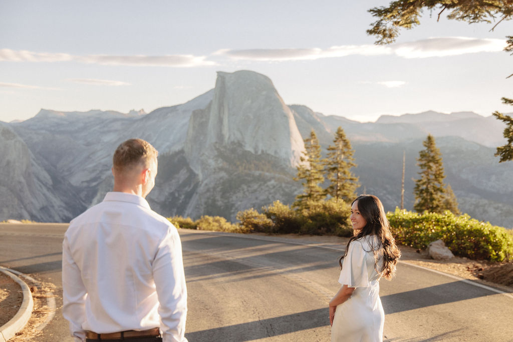 A couple walks hand in hand along a road with Half Dome and the setting sun in the background.