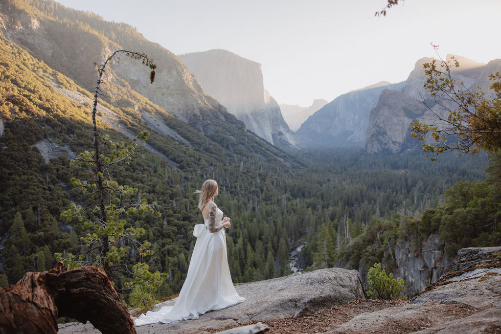 A woman in a white dress stands on a rocky ledge overlooking a valley with distant mountains and forests under a clear sky.