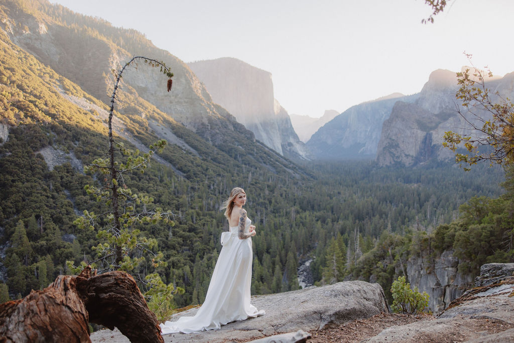 A woman in a white dress stands on a rocky ledge overlooking a valley with distant mountains and forests under a clear sky.