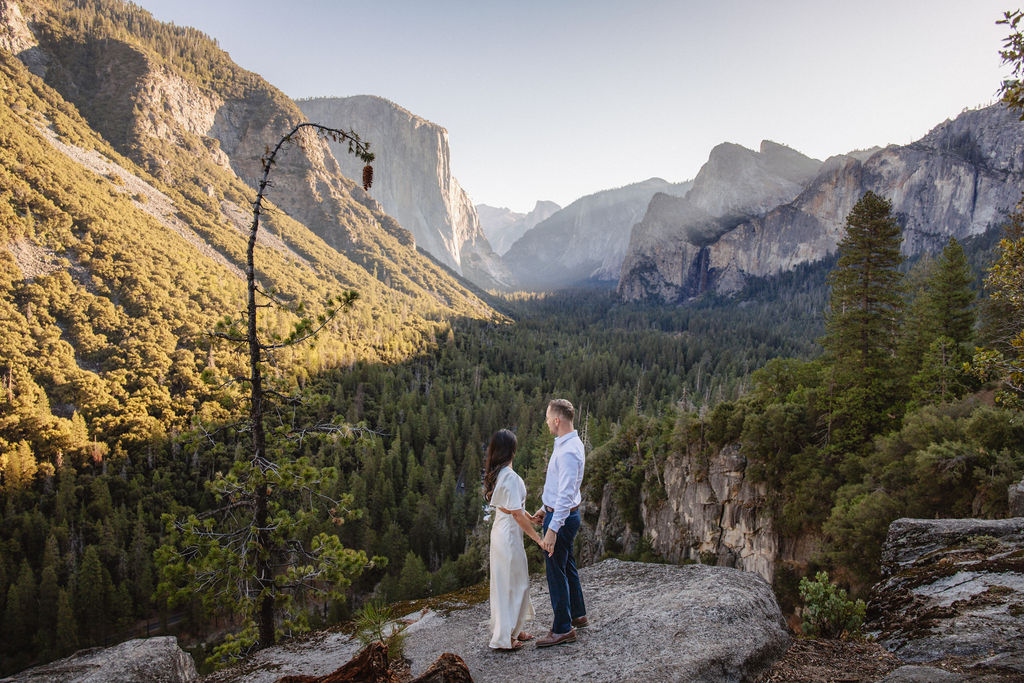 A couple stands on a rocky ledge, overlooking a forested valley with mountains and a clear sky in the background at tunnel view for their yosemite engagement photos