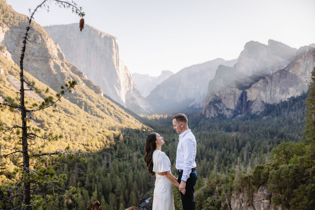 A couple stands on a rocky ledge, overlooking a forested valley with mountains and a clear sky in the background at tunnel view for their yosemite engagement photos