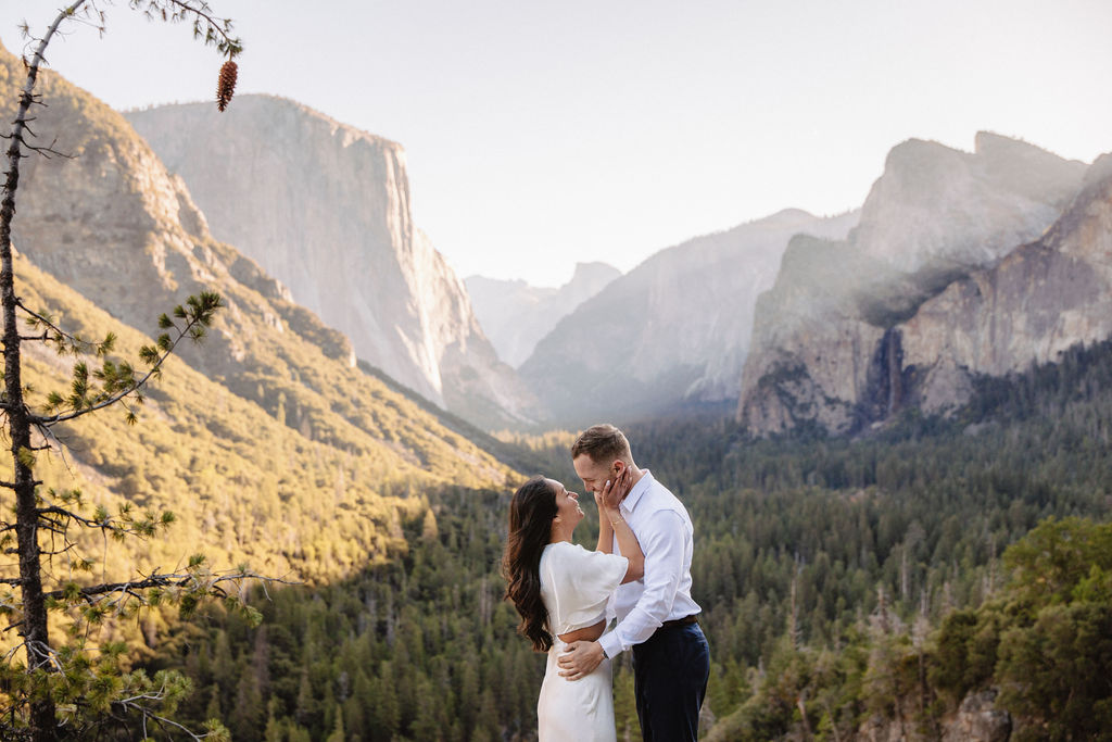 A couple stands on a rocky ledge, overlooking a forested valley with mountains and a clear sky in the background at tunnel view for their yosemite engagement photos