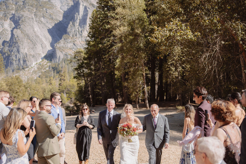 A couple walks hand in hand down an outdoor path during a ceremony, surrounded by a small group of seated and standing guests, with a forest backdrop.