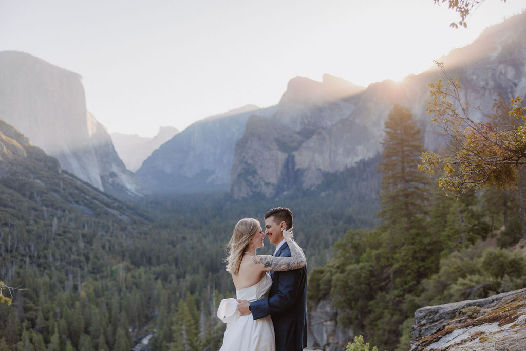 A woman in a white dress stands on a rocky ledge overlooking a valley with distant mountains and forests under a clear sky.