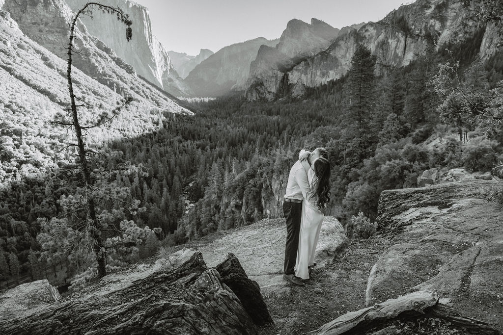 A couple stands on a rocky ledge, overlooking a forested valley with mountains and a clear sky in the background at tunnel view for their yosemite engagement photos