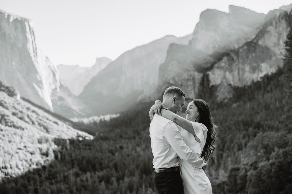 A couple stands on a rocky ledge, overlooking a forested valley with mountains and a clear sky in the background at tunnel view for their yosemite engagement photos