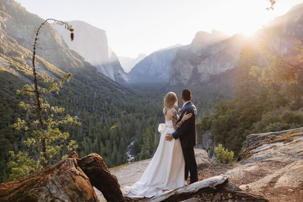 A woman in a white dress stands on a rocky ledge overlooking a valley with distant mountains and forests under a clear sky.