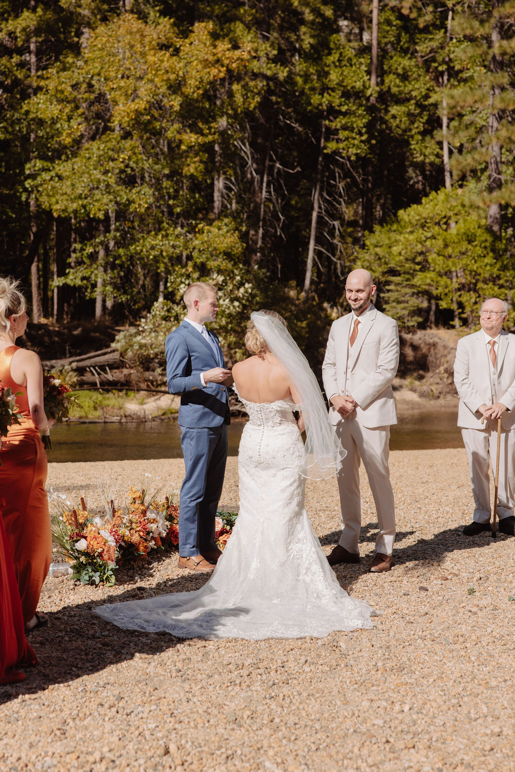 A wedding ceremony outdoors with a rocky mountain backdrop, surrounded by tall pine trees. Guests are seated on either side of the couple and officiant.