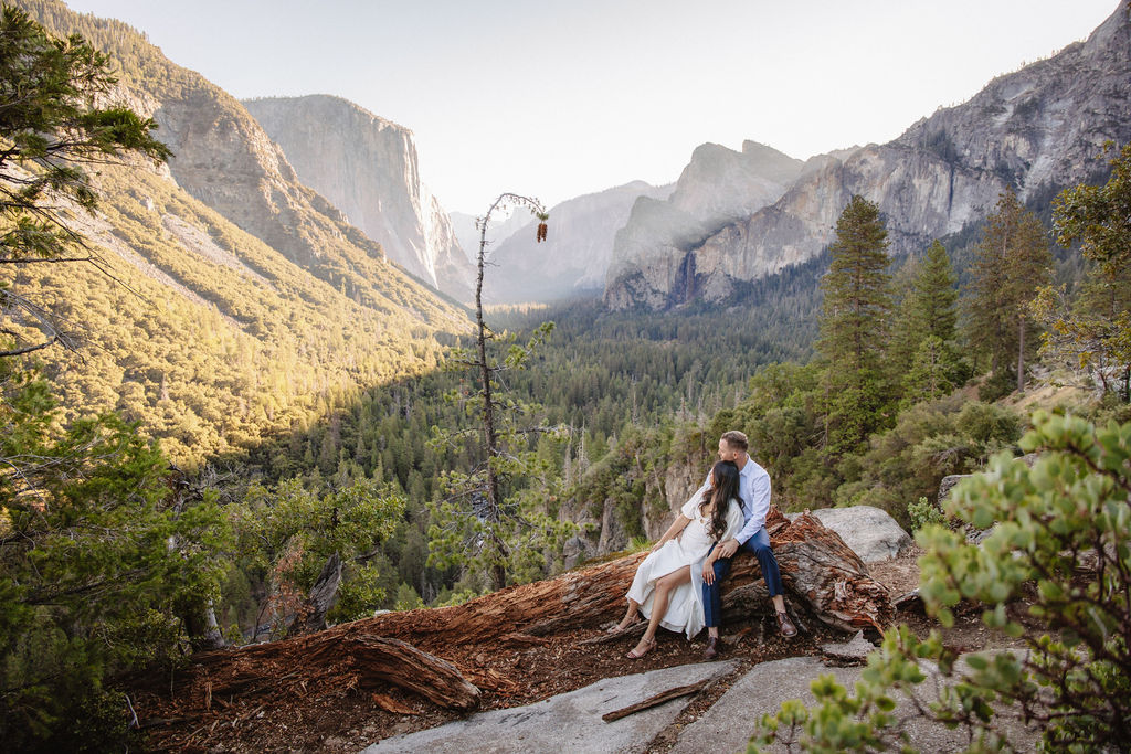 A couple stands on a rocky ledge, overlooking a forested valley with mountains and a clear sky in the background at tunnel view for their yosemite engagement photos