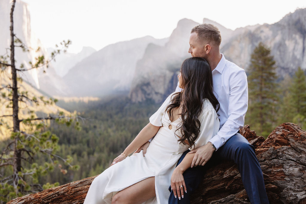 A couple stands on a rocky ledge, overlooking a forested valley with mountains and a clear sky in the background at tunnel view for their yosemite engagement photos