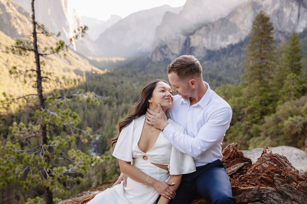 A couple stands on a rocky ledge, overlooking a forested valley with mountains and a clear sky in the background at tunnel view for their yosemite engagement photos