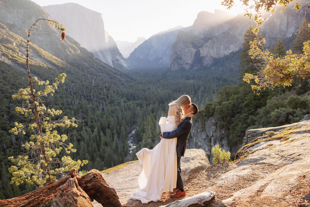 A couple in wedding attire stands on a rocky ledge, embracing with the sun shining behind them. Mountains and trees are visible in the background for an elopement at bridalveil falls in Yosemite