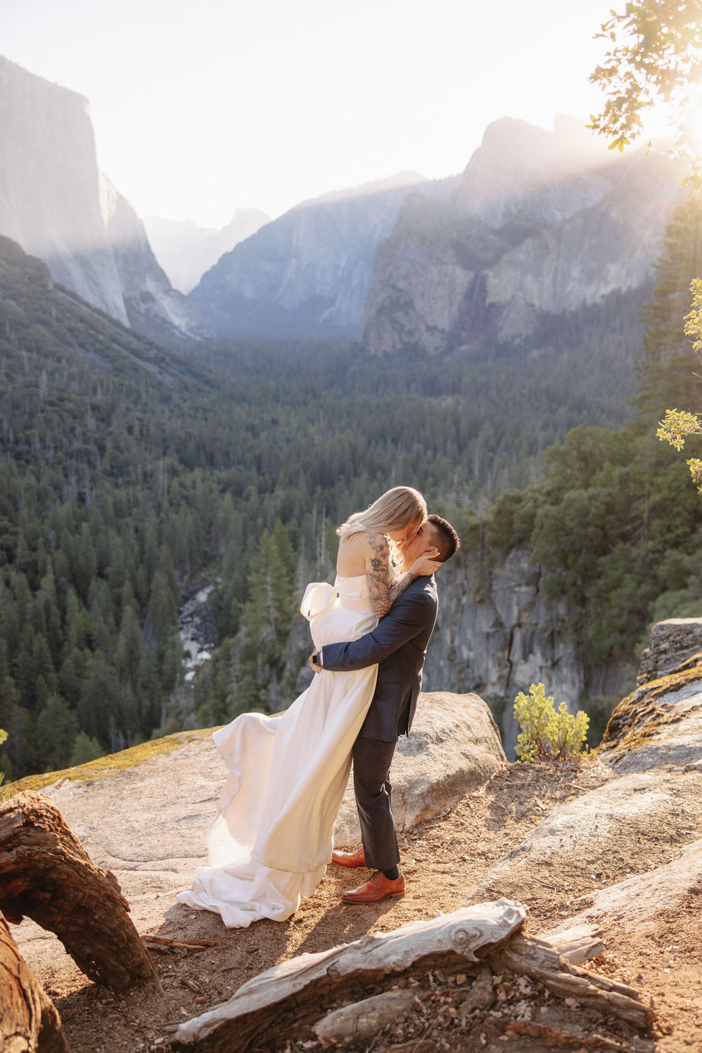 A couple in wedding attire stands on a rocky ledge, embracing with the sun shining behind them. Mountains and trees are visible in the background for an elopement at bridalveil falls in Yosemite