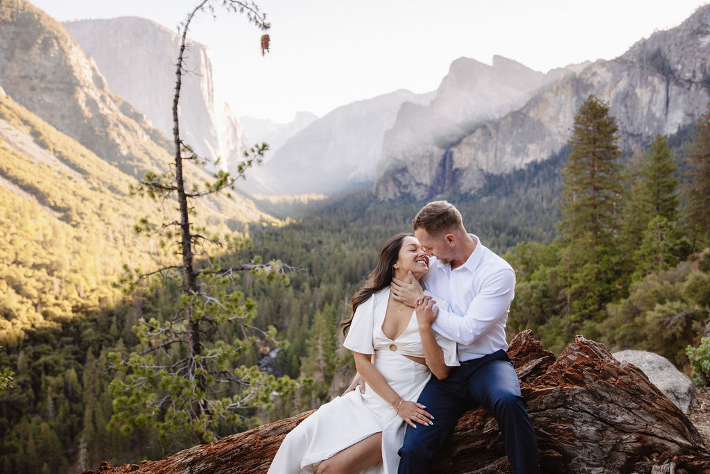 A couple in wedding attire stands on a rocky ledge, embracing with the sun shining behind them. Mountains and trees are visible in the background for an elopement at bridalveil falls in Yosemite