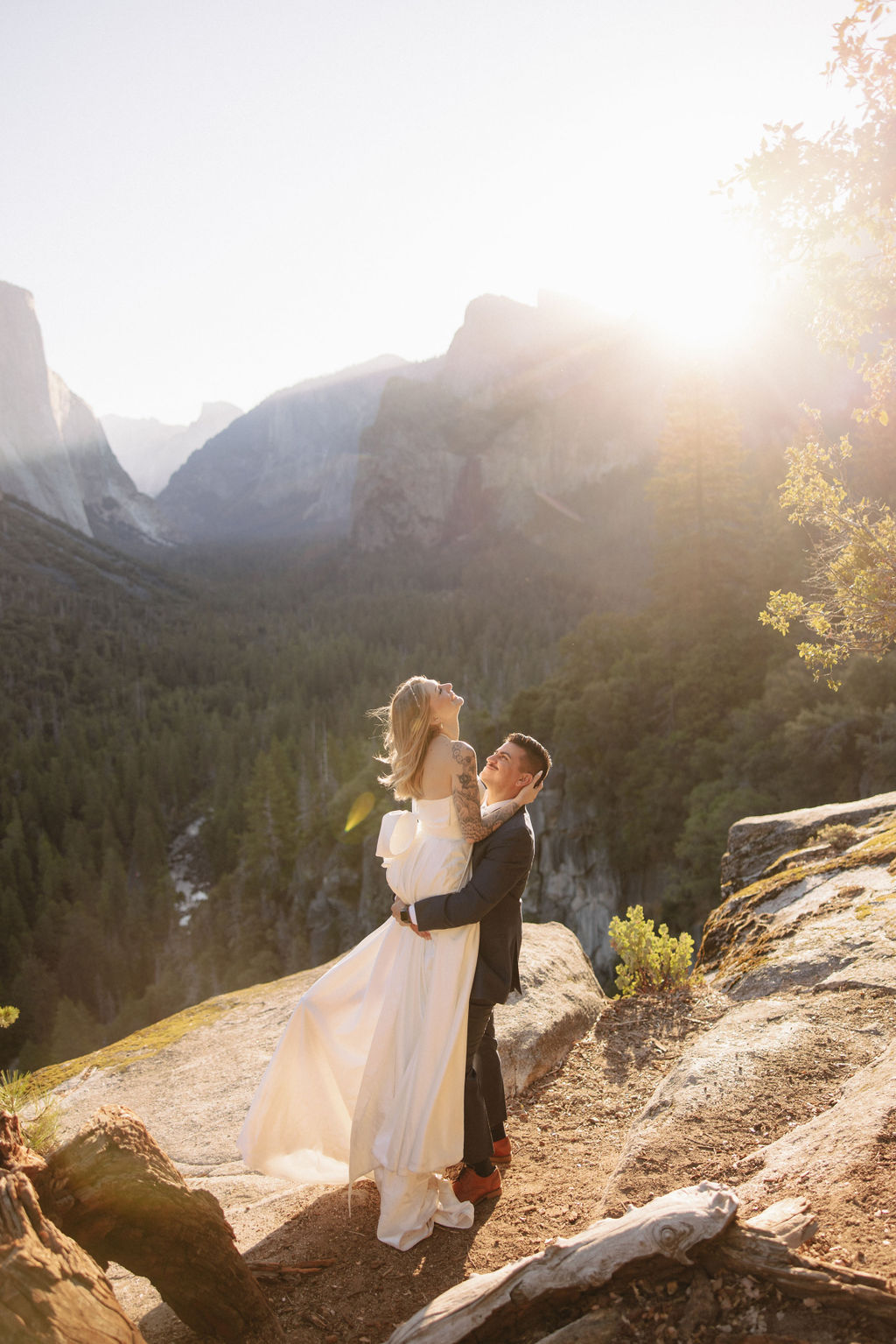 A couple in wedding attire stands on a rocky ledge, embracing with the sun shining behind them. Mountains and trees are visible in the background for an elopement at bridalveil falls in Yosemite