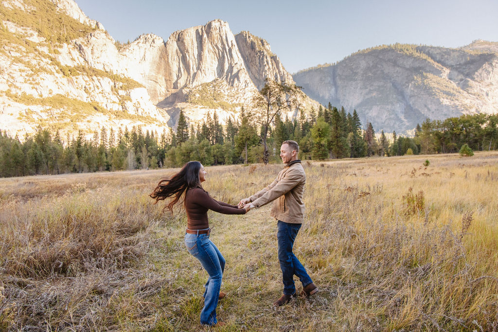 A couple holds hands and spins in a meadow with mountains and trees in the background at swinging bridge meadow for yosemite engaement photos