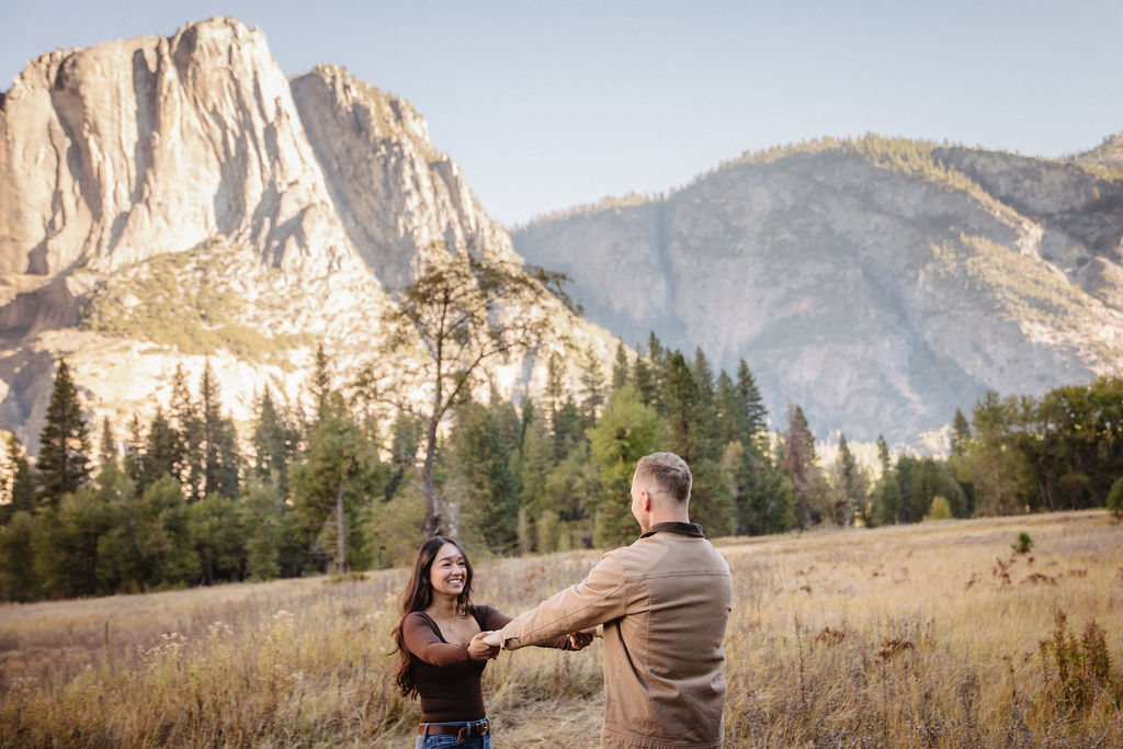 A couple holds hands and spins in a meadow with mountains and trees in the background at swinging bridge meadow for yosemite engaement photos