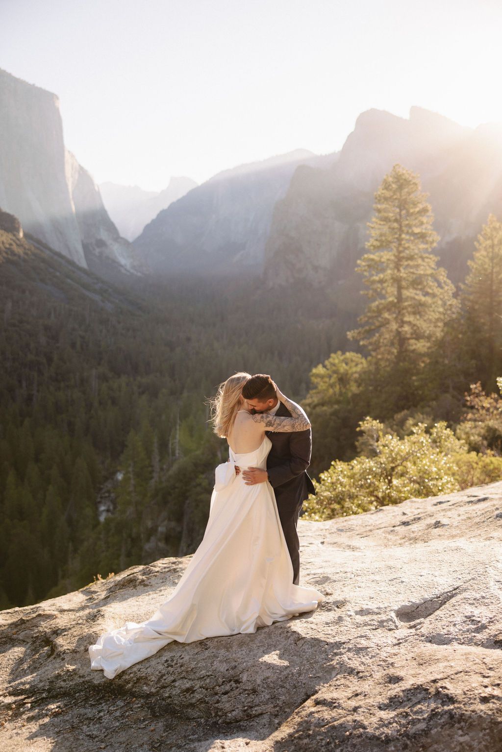 A couple in wedding attire stands on a rocky ledge, embracing with the sun shining behind them. Mountains and trees are visible in the background for an elopement at bridalveil falls in Yosemite