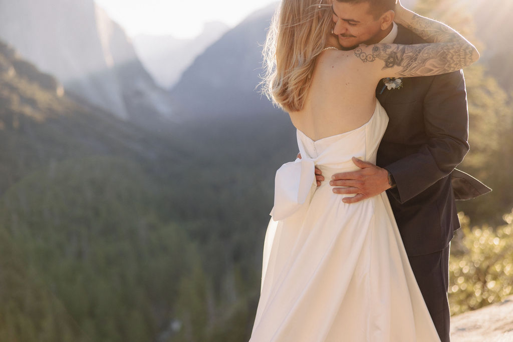 A couple in wedding attire stands on a rocky ledge, embracing with the sun shining behind them. Mountains and trees are visible in the background for an elopement at bridalveil falls in Yosemite