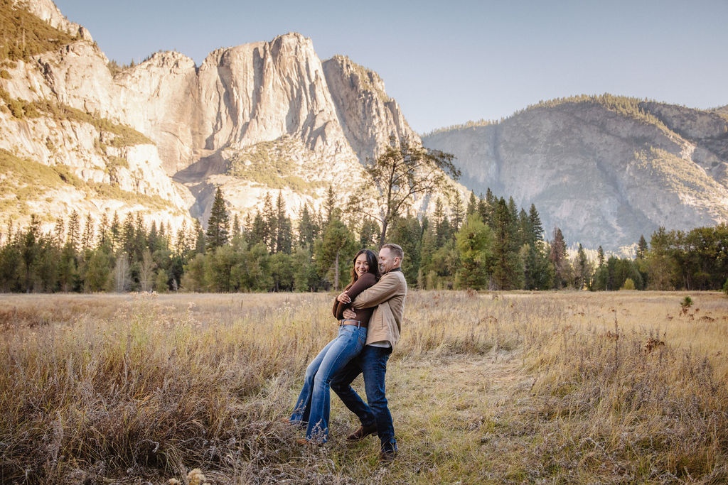 A couple holds hands and spins in a meadow with mountains and trees in the background at swinging bridge meadow for yosemite engaement photos