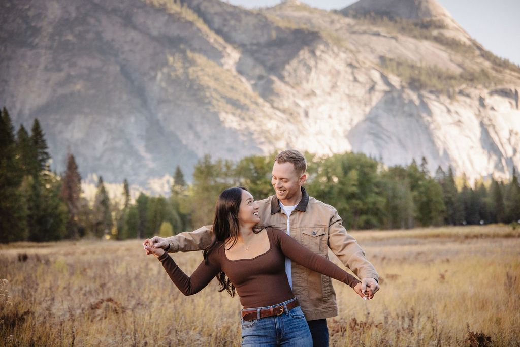 A couple smiles and holds hands in an open field with mountains in the background.