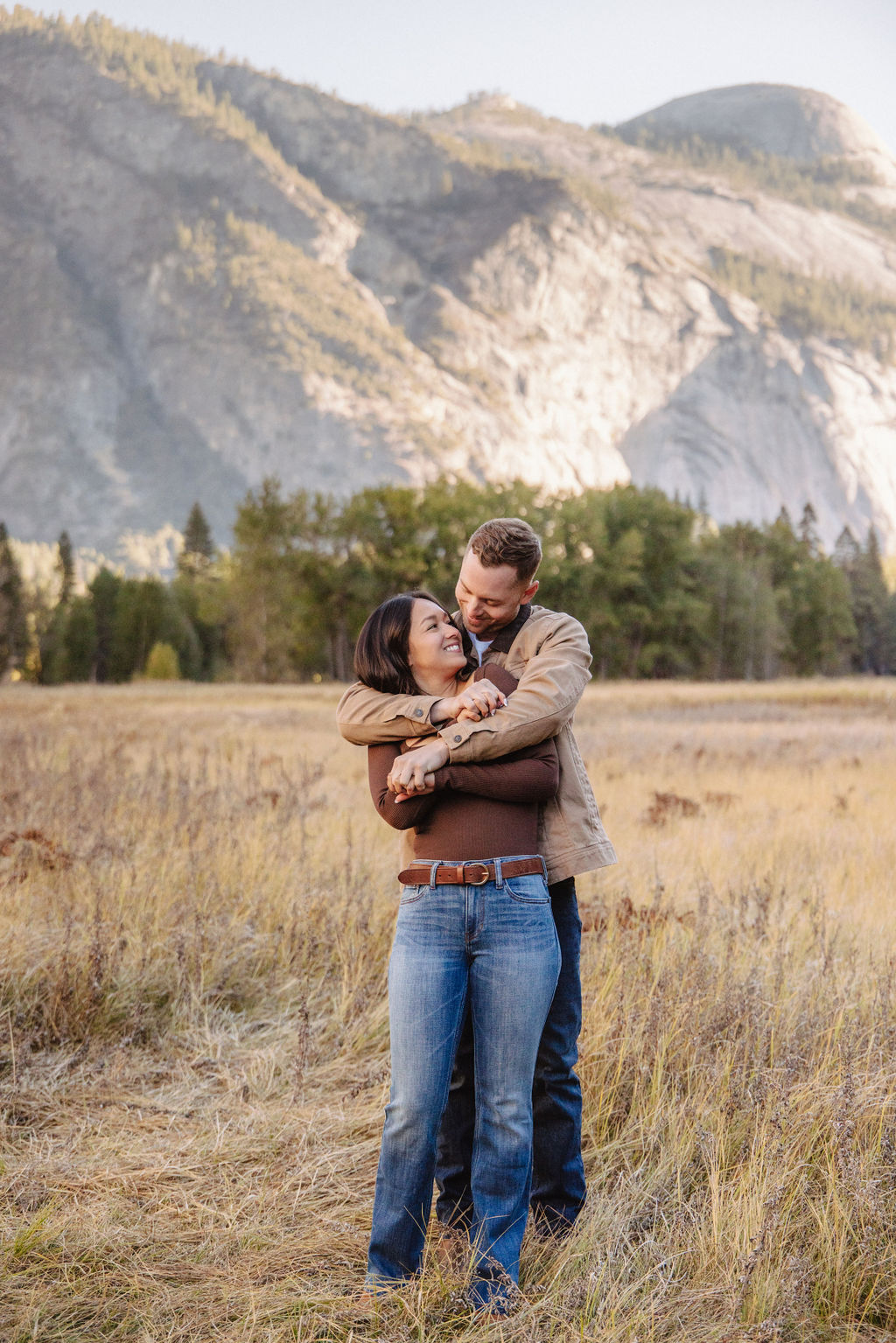 A couple holds hands and spins in a meadow with mountains and trees in the background at swinging bridge meadow for yosemite engaement photos