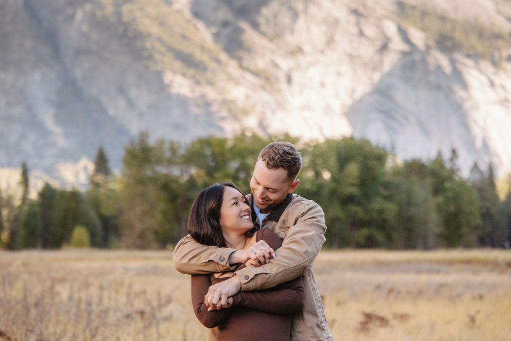 A couple smiles and holds hands in an open field with mountains in the background.