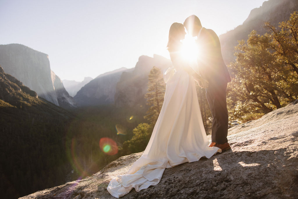 A couple in wedding attire stands on a rocky ledge, embracing with the sun shining behind them. Mountains and trees are visible in the background for an elopement at bridalveil falls in Yosemite