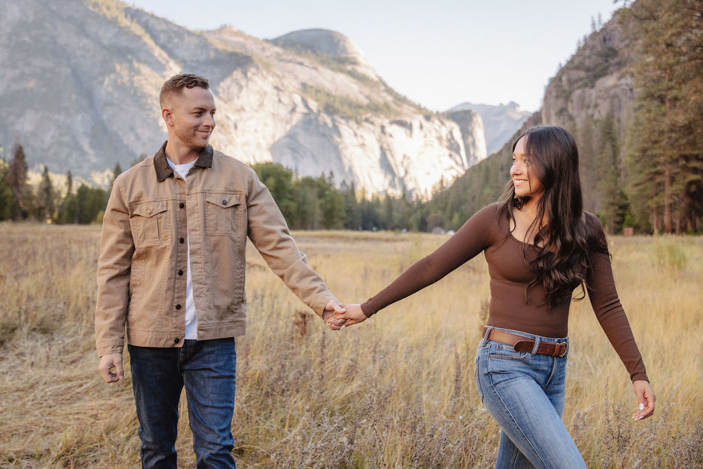 A couple holds hands and spins in a meadow with mountains and trees in the background at swinging bridge meadow for yosemite engaement photos