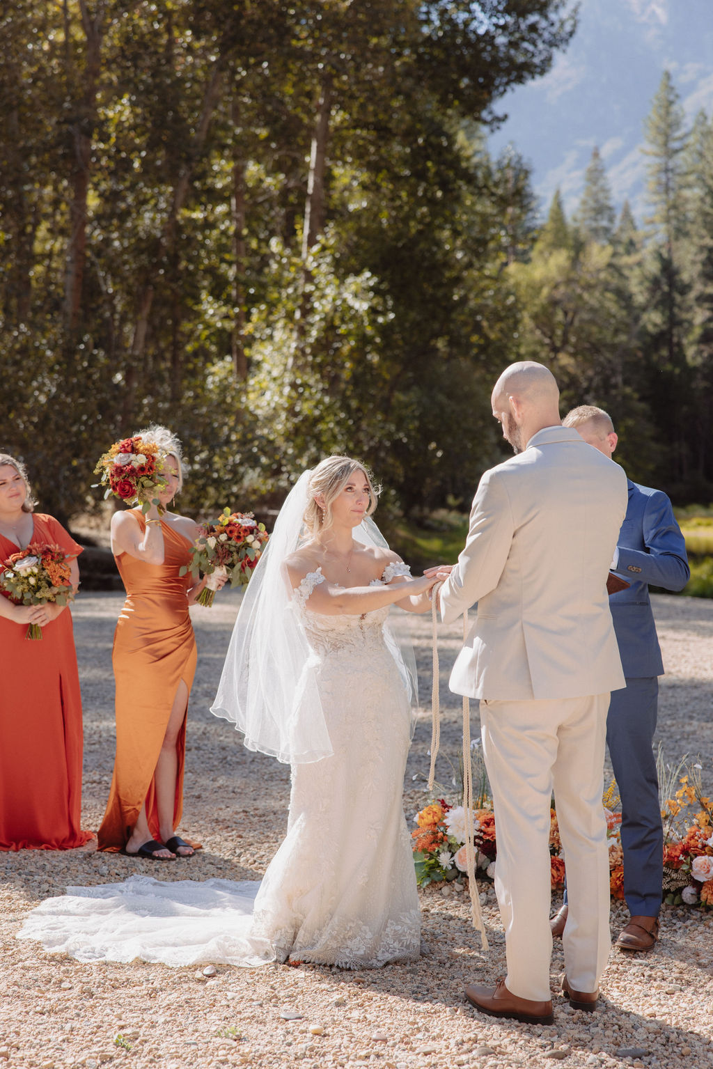 A wedding ceremony outdoors with a rocky mountain backdrop, surrounded by tall pine trees. Guests are seated on either side of the couple and officiant.