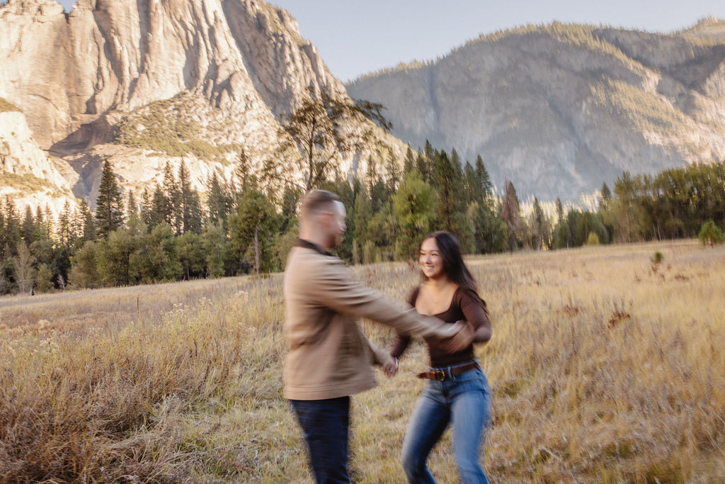 A couple smiles and holds hands in an open field with mountains in the background.